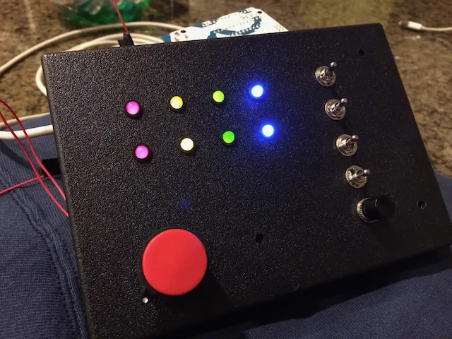A hand-built black control box with glowing coloured LEDs, toggle switches and a big red button
