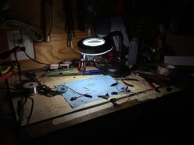 A workbench at night lit by a magnifier lamp, with parts and tools laid out